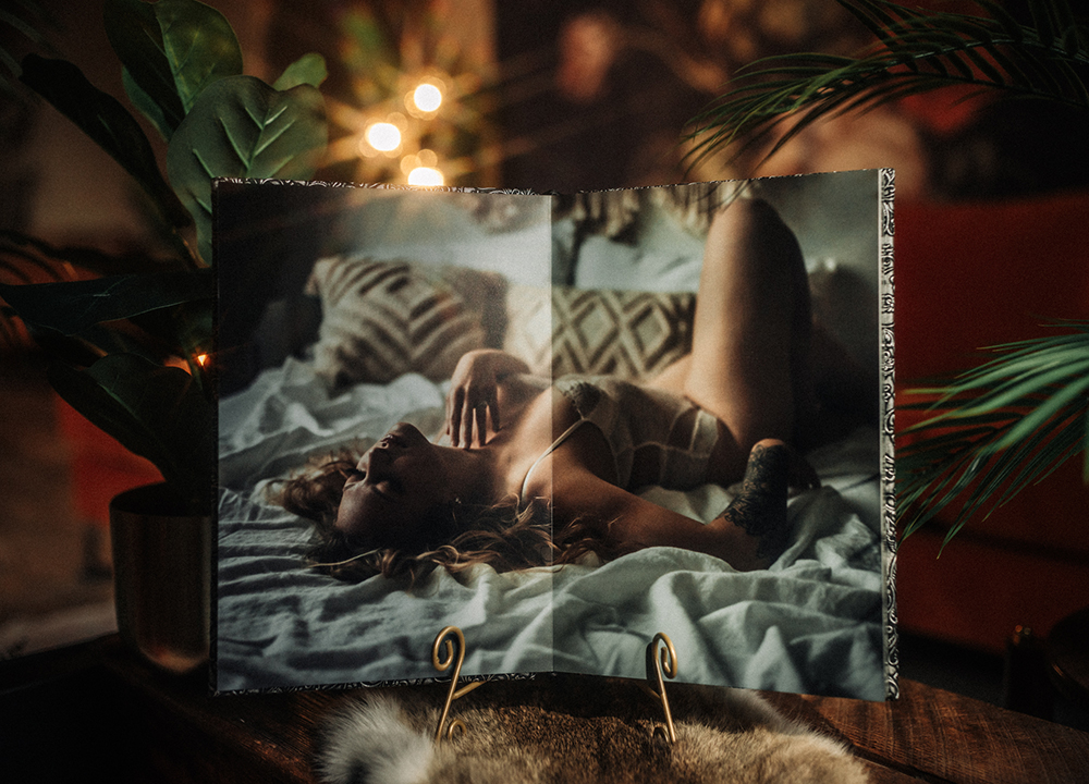 Open luxury boudoir photo album displaying a full spread bedroom boudoir portrait on a decorative stand with soft warm lighting.