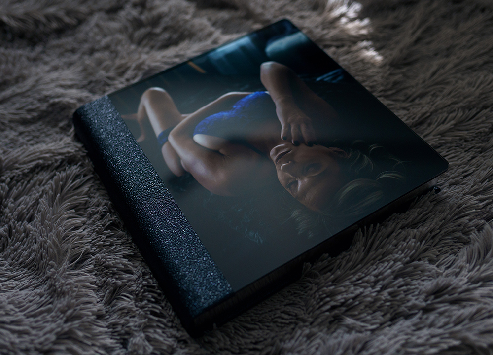 Luxury boudoir photo album with acrylic cover and leather binding displaying a boudoir portrait, resting on a soft textured blanket.