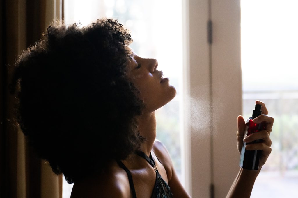 Woman with natural curly hair in black lace lingerie tilting her head back with eyes closed while misting her face near a bright window, during a boudoir session near Houston, Texas.
