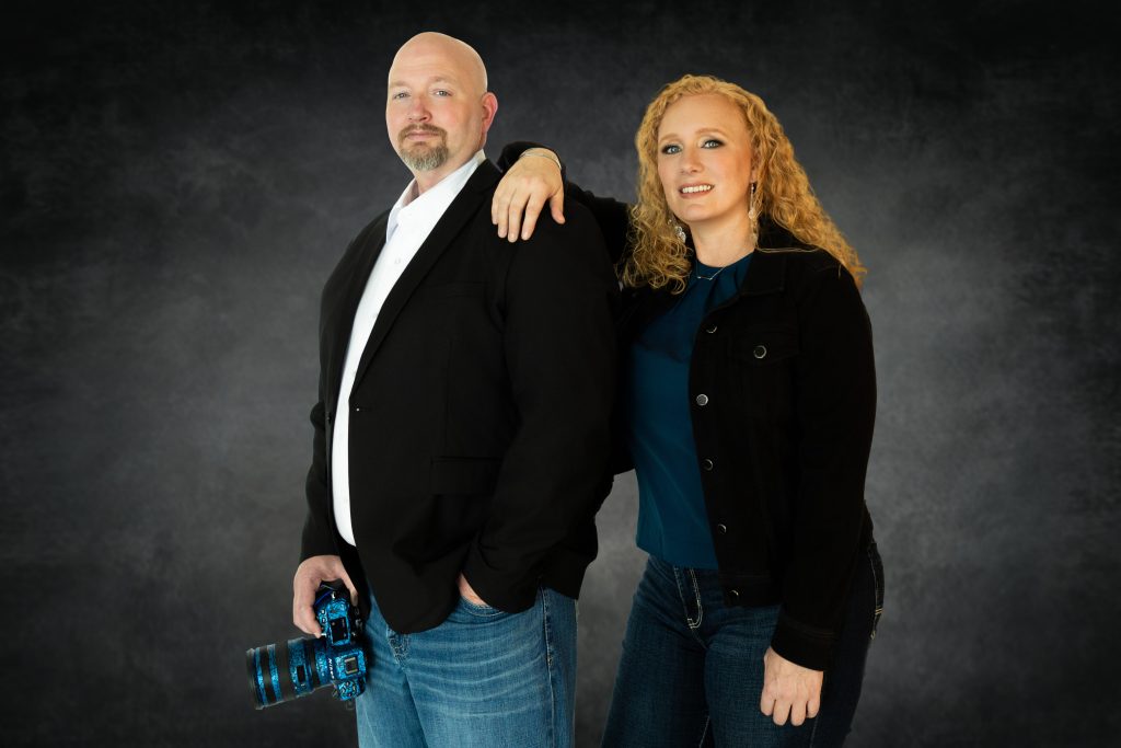 Chris Davis and Heidi Fritsche, owners of Primal Soul Boudoir, stand together in a professional studio portrait holding a camera near Houston, Texas.