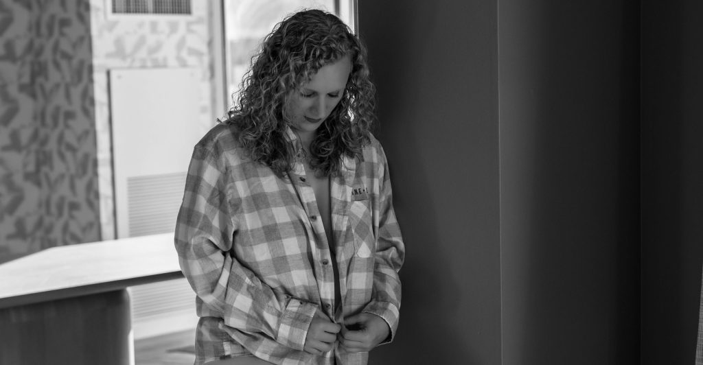 Black and white portrait of a woman wearing an oversized plaid flannel shirt with softly curled hair as she looks down while buttoning the shirt during a boudoir session near Houston, Texas.
