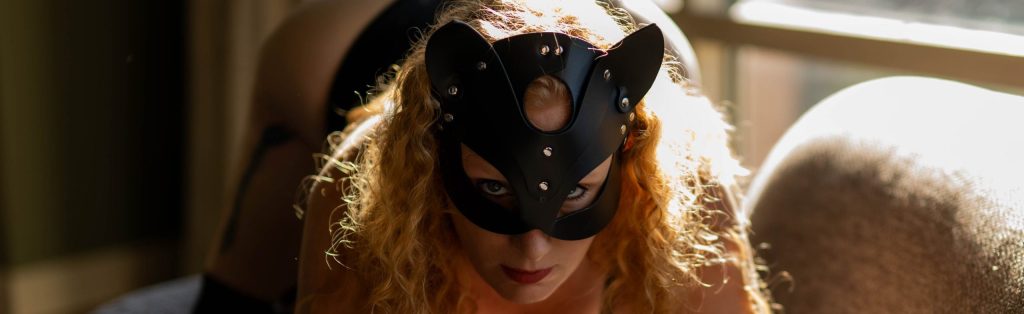 Redheaded woman in a black leather cat mask gazes intently at the camera with dramatic backlit sunlight during a boudoir session near Houston, Texas.