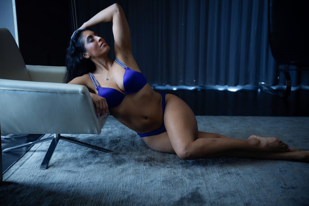 A woman in vibrant royal blue lingerie reclines on the floor beside a white lounge chair with one arm raised above her head in a modern studio setting during a boudoir session near Houston, Texas.