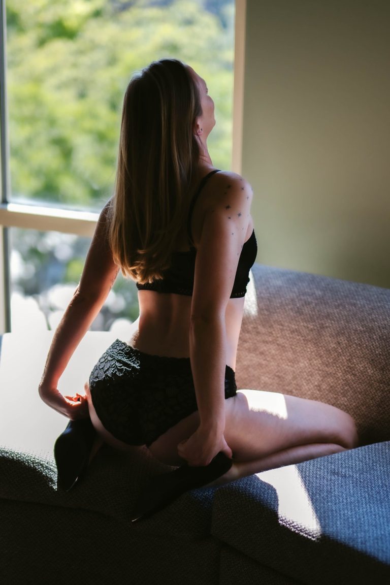 A woman kneels on a sofa in black lace lingerie and high heels, her back turned toward a sunlit window as soft natural light highlights her silhouette during a boudoir session near Houston, Texas.