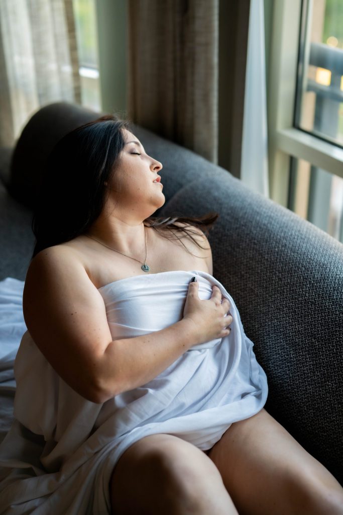 A curvy woman draped in a white sheet sits beside a large window with her eyes closed, gently holding the fabric across her body in soft natural light during a boudoir session near Houston, Texas.