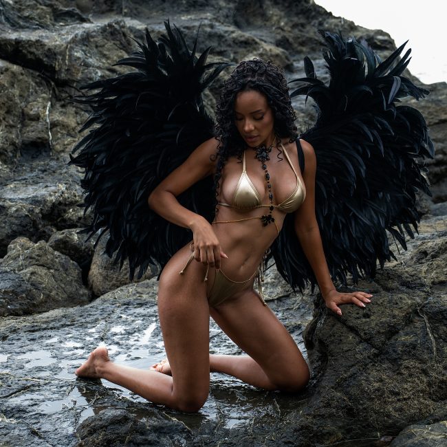 A woman wearing gold lingerie, black crystal body jewelry, and oversized black feather angel wings kneels on ocean rocks during a luxury boudoir session near Houston, Texas.