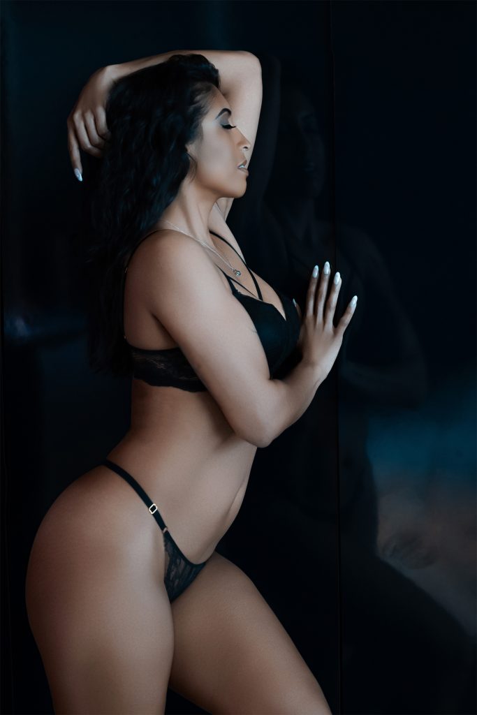 A woman in black lingerie poses in profile against a dark reflective glass wall with one arm raised during a boudoir session in Rosenberg, Texas.
