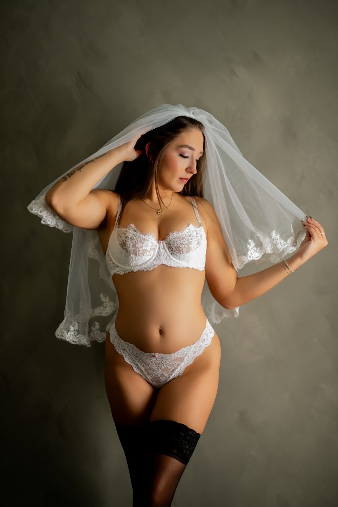 A bride wears white lace lingerie, a delicate bridal veil, and black thigh-high stockings while posing against a neutral studio backdrop during a bridal boudoir session near Houston, Texas.