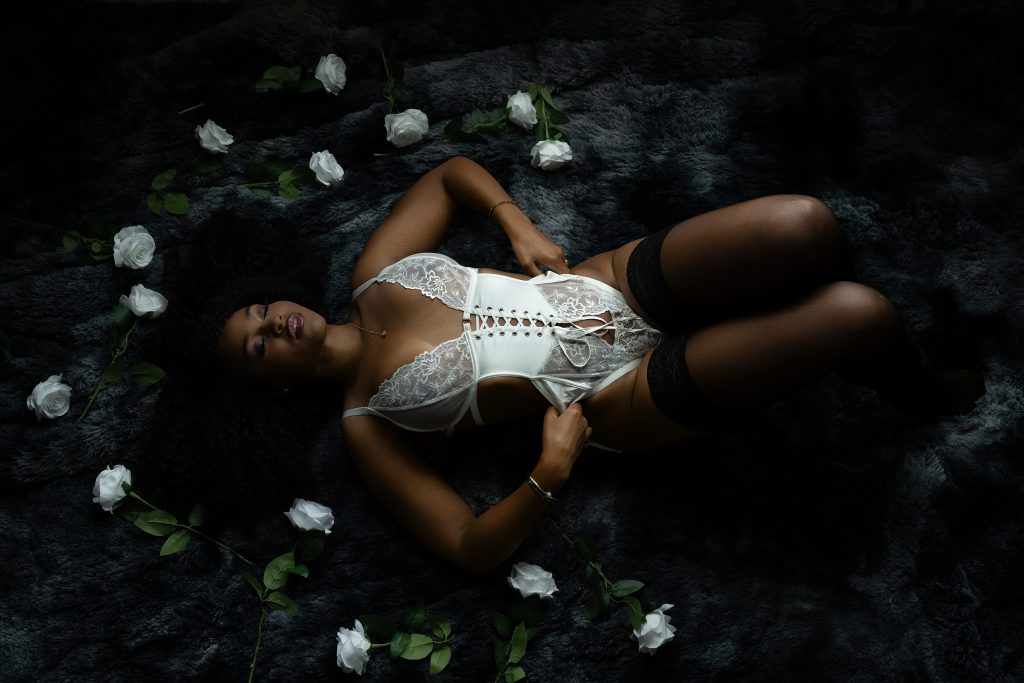 A bride reclines on a dark faux fur rug wearing a white lace corset bodysuit with black thigh-high stockings surrounded by white roses during a boudoir session in Rosenberg, Texas.