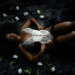 A bride reclines on a dark faux fur rug wearing a white lace corset bodysuit with black thigh-high stockings surrounded by white roses during a boudoir session in Rosenberg, Texas.