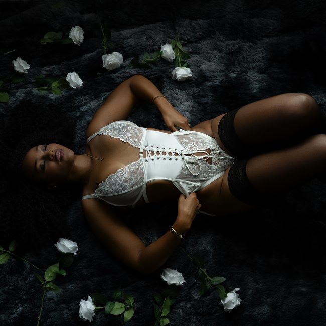 A bride reclines on a dark faux fur rug wearing a white lace corset bodysuit with black thigh-high stockings surrounded by white roses during a boudoir session in Rosenberg, Texas.