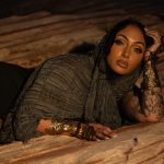 A woman with tattooed arms wears a textured charcoal wrap, black lingerie, and ornate metallic cuffs while reclining on sandstone during an outdoor boudoir session near Houston, Texas.
