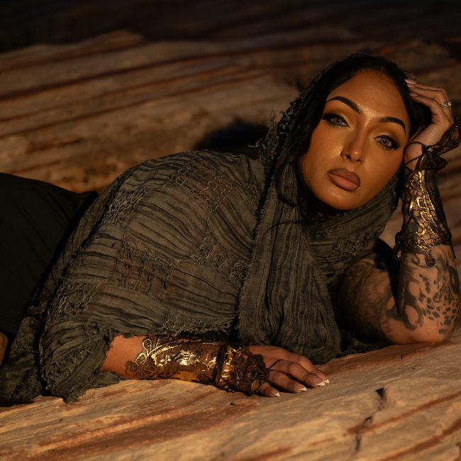 A woman with tattooed arms wears a textured charcoal wrap, black lingerie, and ornate metallic cuffs while reclining on sandstone during an outdoor boudoir session near Houston, Texas.