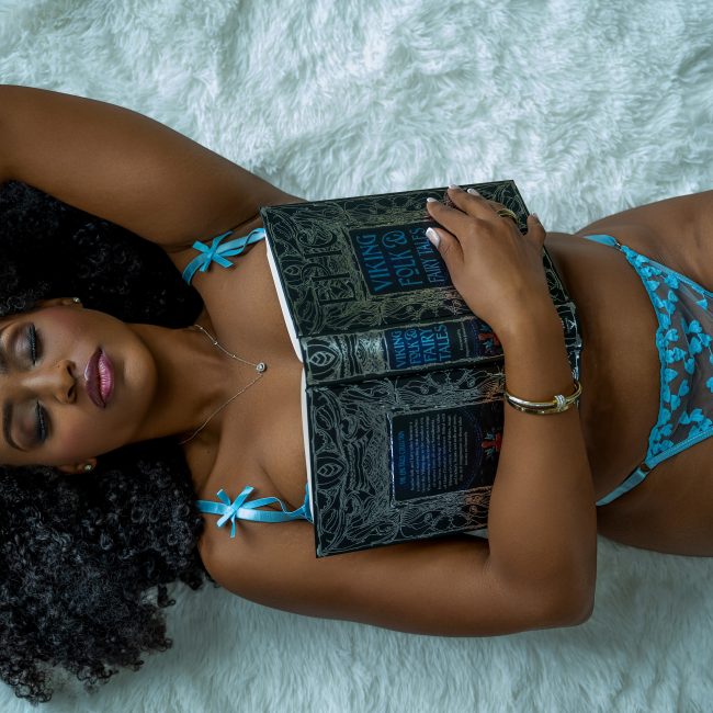 A woman reclines on a white faux fur rug wearing blue embroidered lingerie and black thigh-high stockings while resting an open fairy tale book across her torso during a boudoir session in Rosenberg, Texas.