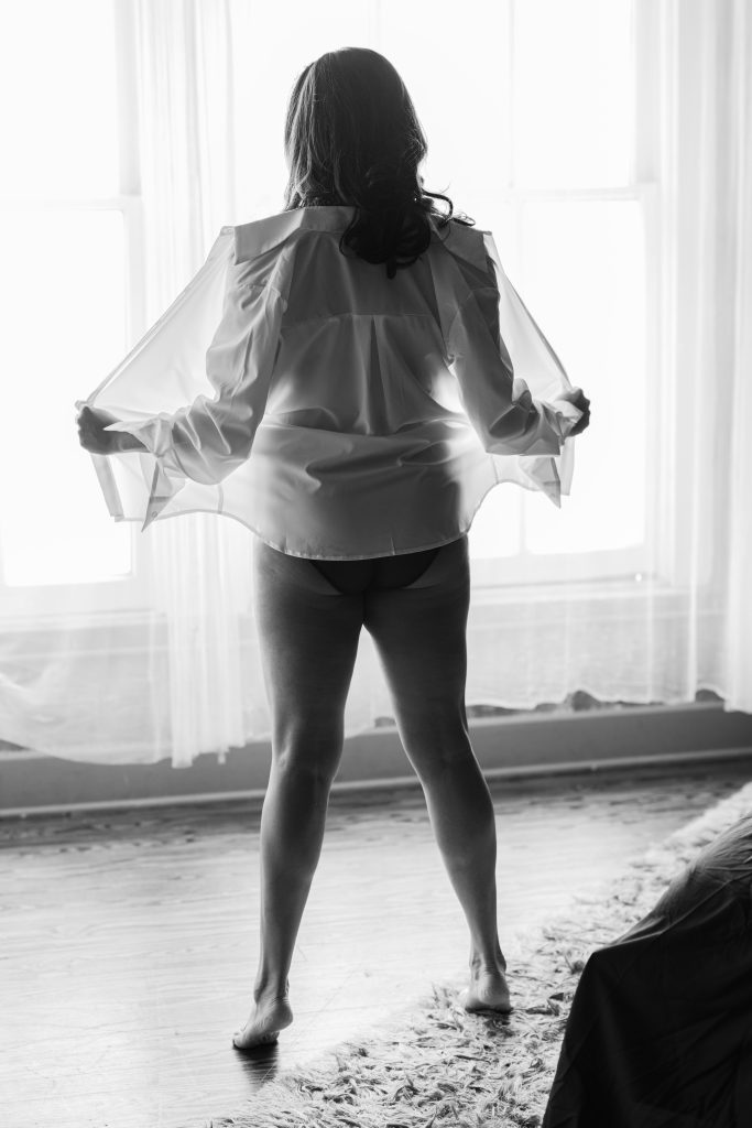 Black and white fine art implied nude portrait featuring a woman standing in front of a sunlit window wearing an open sheer shirt during a luxury boudoir session in Houston.