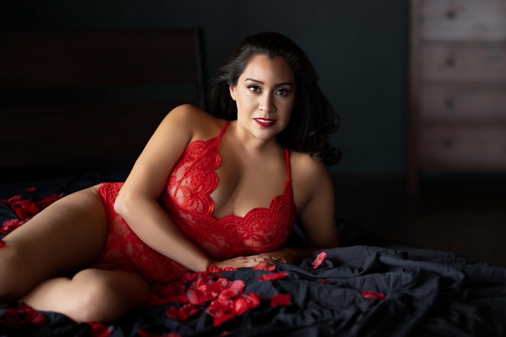 Luxury boudoir portrait of a woman wearing a red lace bodysuit while posing on a bed with rose petals during a Houston boudoir photography session.