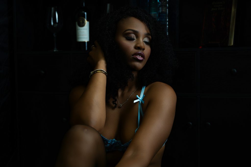 A woman relaxes with her eyes closed wearing delicate blue lingerie in front of a dark wine and book display during a boudoir session in Rosenberg, Texas.