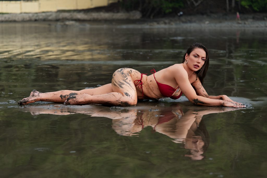 Woman posing in shallow water wearing a red bikini during an adventurous outdoor mud boudoir photography session near Houston, Texas.