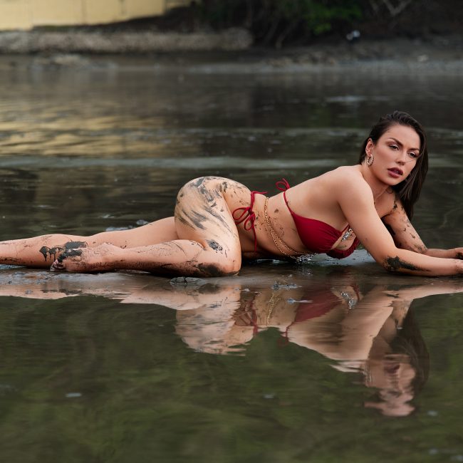 Woman posing in shallow water wearing a red bikini during an adventurous outdoor mud boudoir photography session near Houston, Texas.