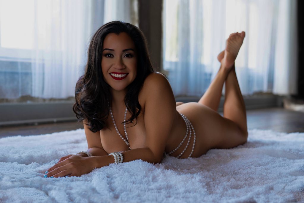 A smiling woman poses on a plush white rug wearing layered pearl jewelry and no visible clothing with her legs raised behind her during a boudoir session in Rosenberg, Texas.