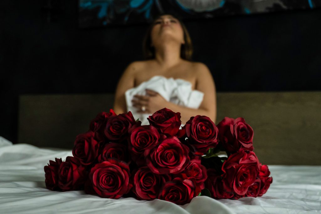 Woman wrapped in white sheets behind a bouquet of red roses during a luxury Valentine's Day boudoir photography session in Houston, Texas.