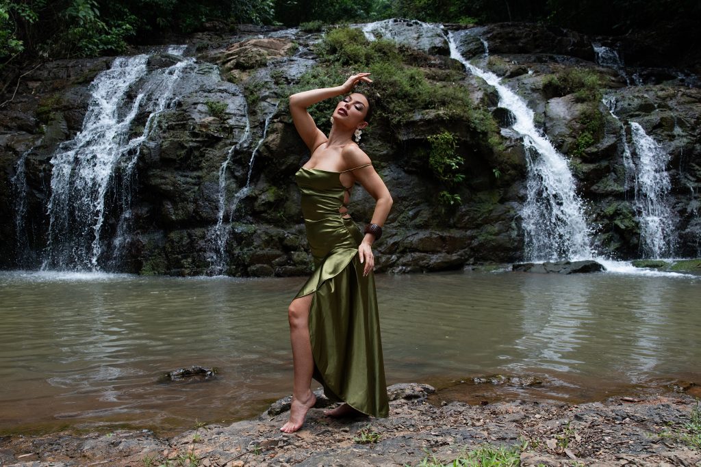 Woman wearing an elegant green gown posing beside a tropical waterfall during an outdoor adventure boudoir and luxury portrait session by Primal Soul Boudoir.