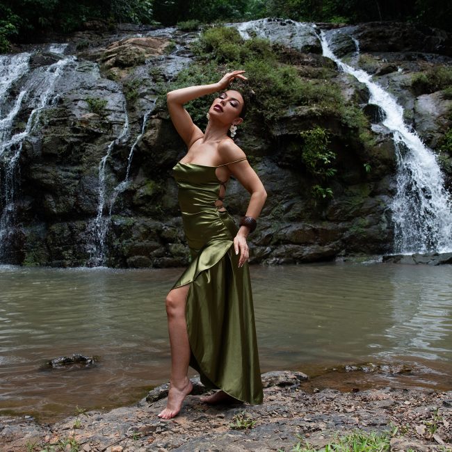 Woman wearing an elegant green gown posing beside a tropical waterfall during an outdoor adventure boudoir and luxury portrait session by Primal Soul Boudoir.