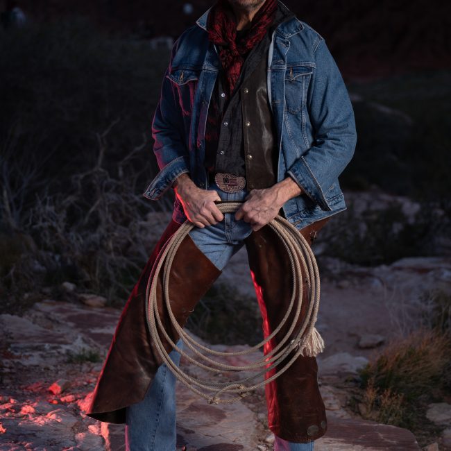 A western cowboy wearing a black hat, denim jacket, leather chaps, and holding a coiled rope stands on sandstone formations at dusk in Red Rock Canyon during a cinematic outdoor portrait session.