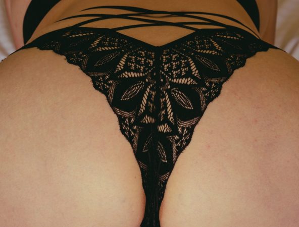 A close-up of a woman lying on a bed wearing black floral lace thong lingerie, highlighting the intricate lace details in soft natural light during a boudoir session near Houston, Texas.