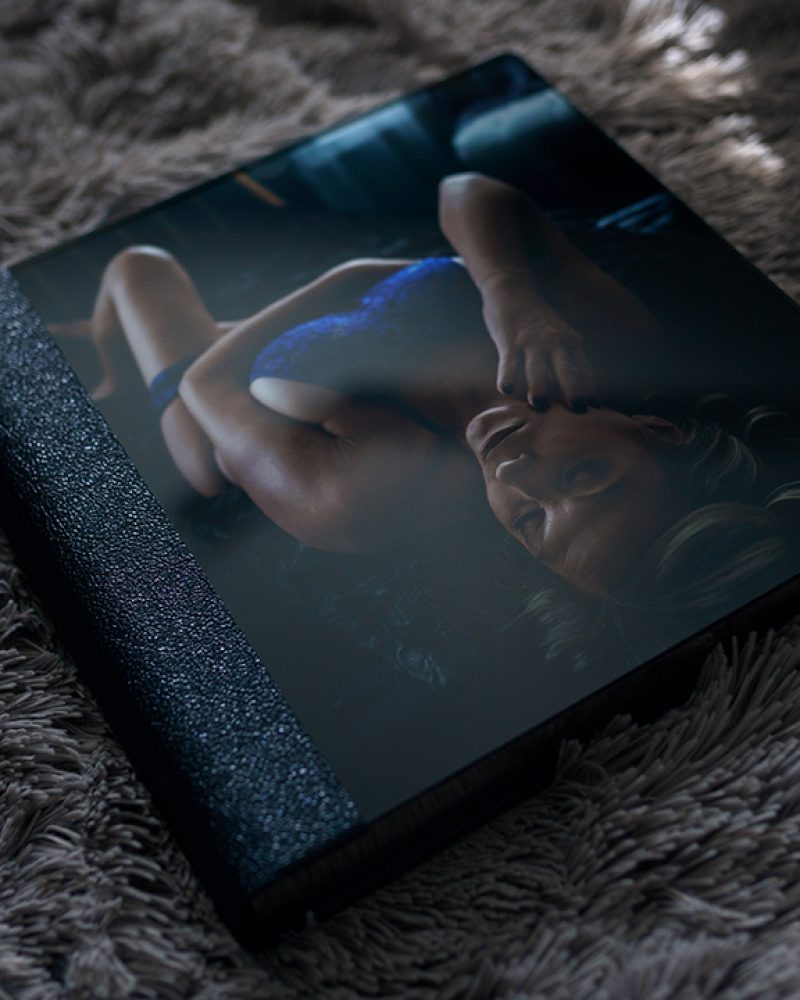 Luxury boudoir photo album with acrylic cover and leather binding displaying a boudoir portrait, resting on a soft textured blanket.